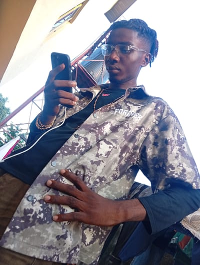 A young Black man, wearing glasses, a black t-shirt, a camouflage jacket with "FOREVER" printed in white on the sleeve, and brown pants, holds a black smartphone in his left hand. He is also wearing a gold chain and a gold bracelet on his left wrist. His right hand is placed on the front of his jacket. The man appears to be looking down at his phone. The background suggests an outdoor setting with a bright sky, some metallic structures, and hints of greenery. The lighting indicates it is daytime, likely sunny. The scene appears to be taking place in Ukukwa, Nigeria.