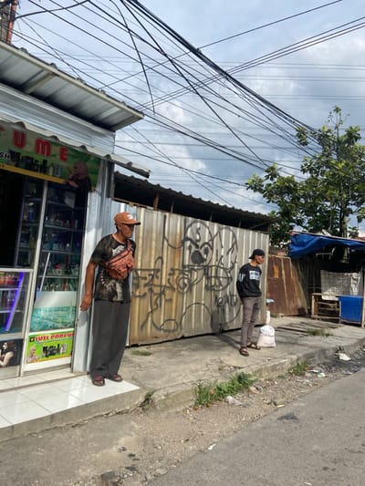 The scene depicts a street corner in Kecamatan Dayeuhkolot, Indonesia (latitude: -6.974, longitude: 107.618). Two adult males are standing on the sidewalk. One man is wearing a brown hat, brown and black patterned shirt, and dark pants. He is standing in front of a storefront with the word "JUME" painted on the side. The storefront has glass shelves filled with various items. The other man is wearing a black and white shirt and black pants. He is standing in front of a large grey metal gate with graffiti. The gate has a large, drawn face on it. Both men are looking away from the camera. There are several power lines strung across the sky. The sidewalk is made of concrete and there are weeds growing on it. The street is paved and empty. There is a small, blue awning to the right of the grey metal gate. It is unclear what is behind the awning, as the scene is obstructed by a large amount of clutter. The scene is a snapshot of a typical Indonesian street scene with a mix of commerce, street life, and graffiti.
