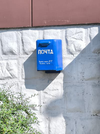 A bright blue mailbox, labeled "ПОЧТА" (POST), is mounted on a textured white brick wall. Below the main label, smaller text reads "Выемка писем с 8^9" and "Выходной Вт, Пн". This translates to "Letter collection from 8-9" and "Days off: Tuesday, Monday". The mailbox casts a shadow on the wall, as does a portion of a dark reddish-brown overhang above it. To the lower left, a portion of a green leafy bush is visible. The scene appears to be outdoors on a sunny day. The context provided places this mailbox in the city of Rybnitsa, Moldova.