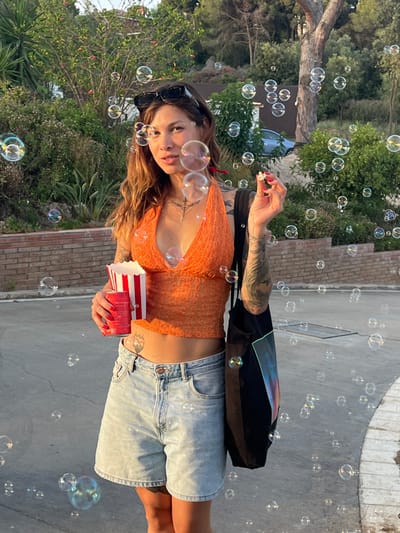 In Sant Vicenç De Montalt, Spain, a young woman stands outdoors with numerous bubbles floating around her. She is wearing a vibrant orange, lace halter top and light-wash denim shorts. In her right hand, she holds a small white object, possibly food, and in her left hand, she holds a red and white striped popcorn container and a red can. She is also carrying a black tote bag slung over her right shoulder. The background features lush greenery, trees, a brick wall, and a paved surface, suggesting an outdoor urban or park setting bathed in the warm glow of sunset.