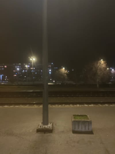 A nighttime urban scene is captured from a high vantage point, possibly a window. Streetlights and building lights illuminate the distant cityscape, which appears blurred. Closer to the viewer, a metal pole stands, affixed to the ground with bolts, with a planter box containing greenery on a concrete surface. A railing or divider is visible between the pole and the distant city lights. The scene is generally dark, with the artificial lights providing the primary source of illumination.