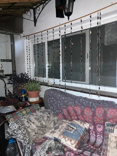 The image depicts an interior scene, likely a covered balcony or porch, with a window as the central element. The window is covered by a mesh screen and adorned with hanging strings of colorful, circular objects, possibly beads or buttons, strung from a wooden rod. A dark, ornate metal bracket is mounted on the wall above the window, supporting an unseen structure. The windowsill is dark and glossy.
In the foreground, a sofa is visible, draped with various patterned textiles and cushions. One cushion has a black and white floral pattern, while others feature geometric and paisley designs in muted colors. A large plastic bottle filled with dark liquid is on the lower left side of the frame. To the left of the sofa, on a table or surface, are two potted plants: one with dark purple, broad leaves and another with thin, green, variegated leaves in a terracotta pot. A can of Raid insect killer is placed next to the purple-leafed plant.
The walls are painted white and show signs of texture. A vintage-style black lantern hangs from the ceiling near the top right corner. The overall impression is one of a lived-in, bohemian-style space.
Regarding the location in Sabadell, Spain, this specific image does not contain any explicit visual cues, such as street signs, landmarks, or architectural styles unique to Sabadell, that would definitively place it within that city. Therefore, I cannot confirm the location as Sabadell, Spain based solely on the provided image.