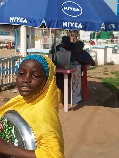 In Garko, Nigeria, a young girl wearing a yellow hijab and blue headscarf is in the foreground, holding a silver tray. Behind her, two men are seated near a table with drinks, under a blue Nivea umbrella. The environment appears to be an outdoor market or roadside stall, with sand and grass visible.