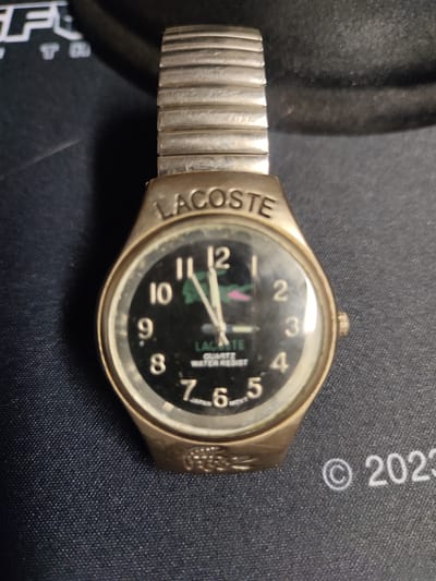 A close-up, top-down shot of a gold-toned, metal-banded wristwatch with a black face is positioned on a dark blue, textured surface. The watch features silver hour markers and hands, with the brand name "LACOSTE" clearly visible above the face and "QUARTZ WATER RESIST" and "JAPAN MOVT" printed on the dial. The watch band is segmented and metallic. To the right of the watch, the year "2023" is visible, also printed on the dark surface. The background is dark and out of focus, but hints of lettering, possibly from a logo, are present in the top left corner. There is no indication of the location being Biliakiemis, Lithuania.
