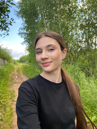 The media file is a portrait of a young woman outdoors. She is facing towards the camera, with her head tilted slightly. She has long, brown hair that is partially visible. Her eyes are open, and she is smiling subtly. She is wearing a black long-sleeved shirt.
The background consists of lush green foliage, suggesting a natural setting like a park or a forest. A dirt path or road is visible in the distance, winding through the greenery. The sky is partly cloudy with patches of blue. The lighting suggests it is daytime, likely late afternoon, with soft, natural light.
The overall mood is relaxed and peaceful. The woman's expression conveys a sense of contentment. There are no visible objects or text in the image. The scene appears to be set in a rural or semi-rural area, consistent with the provided location context of Novyi Ural, Russia. The image is oriented vertically, as if taken with a phone.