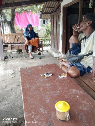 Two people are seated on a wooden bench and a wooden table under a partially enclosed porch. One person is a woman wearing a blue patterned headscarf and orange pants, sitting on a bench with wooden slats. She is looking down. The other person is a man wearing a light-colored shirt, sitting on the bench with his legs outstretched. He is holding a cigarette to his lips and is looking to his right. There is a brown wooden bench and table in front of them. Two small boxes are on the table, and a yellow lidded cup with brown liquid inside is sitting to the right of the table. There is a gray canvas bag on the ground next to the man. The environment is in the shade, and there are trees and plants visible through the open porch. The coordinates place the location in Kecamatan Haurgeulis, Indonesia.