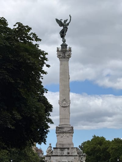 The image shows the Monument aux Girondins, located at the Place des Quinconces in Bordeaux, France. A tall, ornate stone column stands prominently in the center, topped with a bronze statue of a winged figure, often identified as a representation of Liberty. The statue holds a torch in one hand and what appears to be a laurel wreath in the other. The column itself is decorated with bas-reliefs and ornate carvings. To the left of the column, a large, dark green tree with dense foliage dominates the foreground, partially obscuring the monument. In the background, a partly cloudy sky with patches of blue is visible. At the base of the monument, smaller statues and sculptures can be seen, though they are not clearly detailed. The overall scene depicts a monument in an urban park or square, with a focus on the grandeur of the central structure and its allegorical statue.