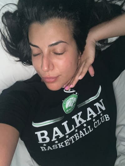 A woman with dark hair is lying in bed, resting her head on her arm. She is wearing a black t-shirt with the words "BALKAN BASKETBALL CLUB" printed on it in white lettering. A logo for the Balkan Basketball Club is visible on the left side of her shirt. The scene appears to be an indoor setting, likely a bedroom. There is no specific information in the image to indicate the location within the city of North Charleston, United States of America.