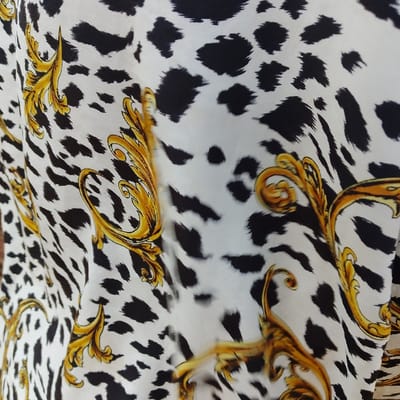 The image displays a close-up of a textile exhibiting a patterned design. The primary pattern consists of irregular black blotches on a white background, consistent with an animal print resembling cowhide or dalmatian spots. Interspersed throughout this black and white pattern are ornate, curvilinear golden-yellow motifs, indicative of baroque-style scrollwork or acanthus leaves. The fabric shows minor folds and creases, suggesting it is draped or loosely arranged. No individuals, other objects, or discernible environmental features are present within the frame. The content of the image itself does not provide any visual information to confirm a specific location within Estômbar e Parchal, Portugal.