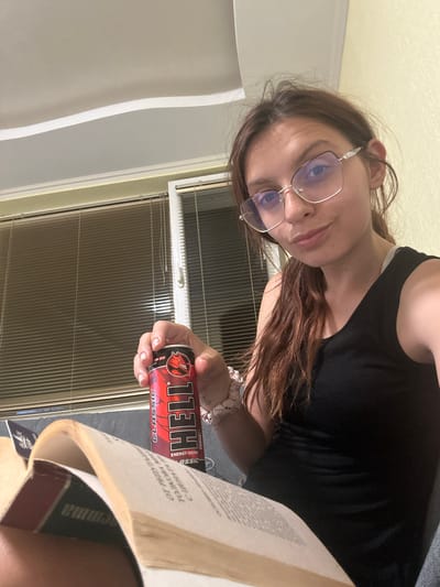 A young woman is sitting indoors, holding a can of "Hell" energy drink and a book. She is wearing glasses and a black tank top. The room has blinds on the window and a ceiling light. The image is taken from a close-up perspective, focusing on the woman and the objects she is holding. The context of Tryavna, Bulgaria is not directly evident from the visual information in the image.