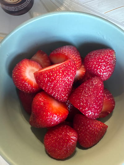 This is a close-up, top-down shot of a light blue bowl filled with fresh, sliced strawberries. The strawberries are bright red, plump, and have visible seeds. Some are whole, while others are halved or quartered, revealing their white interiors and red flesh. The bowl is placed on a surface with a light-colored, textured pattern, possibly a tablecloth or countertop.
To the left of the bowl, a portion of a dark circular object with text is visible. The text is in Italian and appears to be describing a food product, mentioning "biscotti con cuore cremoso" (cookies with a creamy heart) and "cuore onctueux" (creamy heart).
The lighting suggests an indoor setting, possibly during the daytime. There are no people visible in the image. The scene is simple and focuses on the fresh fruit, evoking a sense of healthy eating or a light snack. The overall impression is one of freshness and natural simplicity.