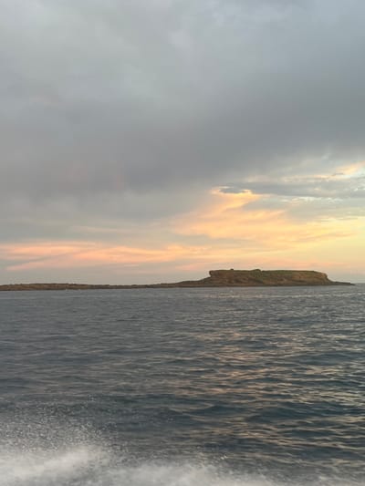 The image displays a marine scene featuring Big Island, located off the coast of Port Kembla, Australia. The foreground shows a white foam wake indicative of a moving vessel, originating from the lower-left corner. The ocean water is dark blue-grey with surface ripples. Big Island, a rocky landmass characterized by a relatively flat top and eroded sides, occupies the midground, extending horizontally across the frame. Sparse green vegetation is visible on the island's upper surface. The sky above is predominantly overcast, with dense grey cloud cover dominating the upper two-thirds. Below this, closer to the horizon, the clouds are illuminated with natural hues of orange, pink, and yellow. No human figures or discernible man-made structures are present within the frame.
