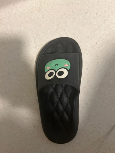 A black slipper with a cartoon frog face is lying on a light-colored tiled floor. The frog's face is green with large white and black eyes and pink cheeks. The slipper is open-toed and has a quilted pattern on the footbed. The floor is made of small, speckled tiles. There is no indication of the slipper's location in Tromsø, Norway.