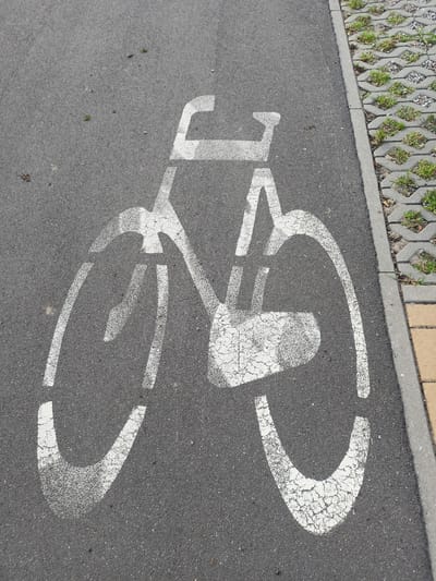 A white bicycle symbol is stenciled on a dark gray asphalt surface, indicating a bike lane. The symbol is comprised of a frame, two wheels, and handlebars. The paint is worn and cracked in places, suggesting age and use. To the right of the asphalt, there is a concrete border, followed by an area of grey concrete pavers with small patches of green grass growing between them. A strip of yellow paving stones is visible at the far right edge of the image. This scene is likely located on a street or path in Grudziądz, Poland, designed for bicycle traffic.