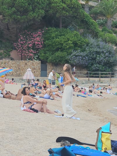 The image depicts a sunny day at a crowded beach in Blanes, Spain, likely Cala Giverola or a similar cove along the Costa Brava. In the foreground, a woman with braided hair is standing and appears to be mid-motion, perhaps dancing or posing, wearing light-colored wide-leg pants and a bikini top. Behind her, numerous people are relaxing on the sand, sunbathing, and socializing. Some are lying on towels, while others are sitting or leaning against beach bags. Beach umbrellas and towels of various colors are scattered across the sand.
The background features a lush, green hillside covered in trees and shrubs. Pink flowering bushes and purple-hued foliage are visible, adding color to the natural landscape. A stone wall runs along the base of the hill, and a wooden fence demarcates the edge of the beach area. A few palm trees are also present. The sea is visible in the far right of the image, with gentle waves lapping at the shore. The overall atmosphere is one of a popular tourist destination, with many people enjoying a leisurely day at the beach. In the lower right corner, beach equipment including a paddleboard and bags are visible.