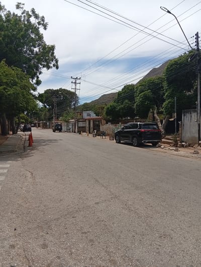 The image depicts a street in Atamo Norte, Venezuela. The road is paved but shows signs of wear. On the left side of the road, there is a sidewalk and several trees, with an orange traffic cone near the curb. Further down the street, there is a dark-colored SUV parked on the right side. Across from the SUV, there is a building and various objects such as a small table and chairs. In the distance, another dark-colored vehicle is visible. Overhead, power lines crisscross the sky. There is a mountain range and a partly cloudy sky.