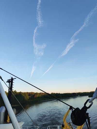 An outdoor, eye-level shot captures a peaceful river scene from the stern of a moving boat at Volokov Most, Russia. The viewpoint looks across the water towards a forested bank under a clear, bright blue sky.
In the immediate foreground, various industrial elements of the boat are visible. To the left, a thick white support structure or mast rises, accompanied by a black, utility-laden pole with multiple attachments and taut ropes. These ropes stretch diagonally across the frame, leading towards a large, dark, rusty pulley or block assembly secured by a shackle on the far right. Portions of yellow machinery are also visible below the ropes. The boat's movement is indicated by the gentle wake trailing in the water.
The middle ground is dominated by a wide body of water, likely a river or canal, reflecting the sky and the surrounding greenery. The water surface shows subtle ripples. On the distant bank, a dense line of lush green trees forms a continuous forest, appearing darker in some areas, possibly due to shadows or different tree types. A tall, slender communication tower or similar structure is faintly visible through the trees on the far bank, towards the right.
The sky occupies the upper two-thirds of the image, showcasing a vibrant blue hue that lightens slightly towards the horizon, suggesting late afternoon or early evening light. Several distinct white contrails streak across the sky; one on the right is long and linear, while another on the left forms a more complex, undulating, almost S-shaped pattern. There is no visible text in the image.