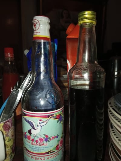 The scene is set indoors in Melakudikadu, India, under dim lighting. The foreground features a collection of domestic items resting on a dark, flat surface.
A white ceramic mug with a floral pattern of pink roses and green leaves is positioned on the left, holding several metal utensils, including spoons and knives. To its right stands a clear glass bottle, approximately three-quarters full with a dark, purplish liquid. Its neck is sealed with a crumpled white, red, and yellow foil cap. The bottle's lower half bears a label depicting a white heron or crane flying over pink and purple flowers, with "Cap BANGAU" prominently displayed, along with "PT. Bangau Sari Mega Jaya Tangerang 15129 INDONESIA" and "PISANG AMBON." An embossed floral design is visible on the bottle's shoulder.
To the right of the first bottle, a second clear glass bottle with a gold-colored screw cap contains a dark, opaque liquid, filling approximately two-thirds of its volume. The name "Mariam" is embossed on its shoulder.
In the mid-ground, a partially visible red bottle with a red cap containing a red liquid is to the left, behind the utensil holder. Behind the prominent left bottle, a blue container is partially visible. Between and behind the two main bottles, an orange carton with an orange cap and a partial white circular logo is present. To the far right, a stack of white and brown patterned plates is visible. The background is dark and indistinct, with a faint framed image or painting depicting a person, possibly a woman, discernible. There are no explicit actions or interactions depicted.