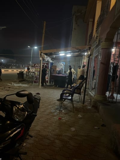 A nighttime view of a street scene shows an outdoor eatery with several people gathered around tables. A motorcycle is parked in the foreground. The area is illuminated by bright overhead lights, and a street lamp is also visible in the distance. The surrounding buildings appear to be multi-story structures, and the street is paved with patterned stones.