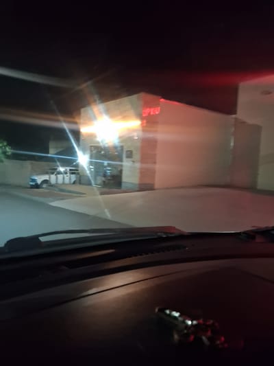The image depicts a night scene viewed from inside a vehicle, looking out at a building. The building has a light-colored facade with illuminated signage that appears to read "OPEN". In front of the building, a white van or SUV is parked. The foreground of the image shows the interior of the car, including the dashboard and windshield. The scene is dimly lit by artificial lights from the building and streetlights, creating lens flares and a slightly blurry effect. There is no identifiable specific location in Yeghegnadzor, Armenia, visible in the image.