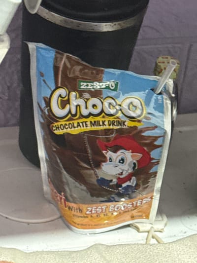 A chocolate milk pouch is shown in the foreground, with a cartoon cow character wearing a red bandana on the front. The pouch is blue and brown, with the word "Choco" in large yellow letters. Text on the pouch includes "CHOCOLATE MILK DRINK" and "WITH ZEST BOOSTERS". The pouch is lying on a white surface, with a dark object to the left that appears to be a thermos or container. The background is out of focus and appears to be a shelf or wall. The lighting suggests an indoor setting, possibly a refrigerator or pantry. The orientation of the pouch and the visible text suggest it is a product commonly found in households or convenience stores in Manila, Philippines. The overall impression is that of a child-friendly beverage.