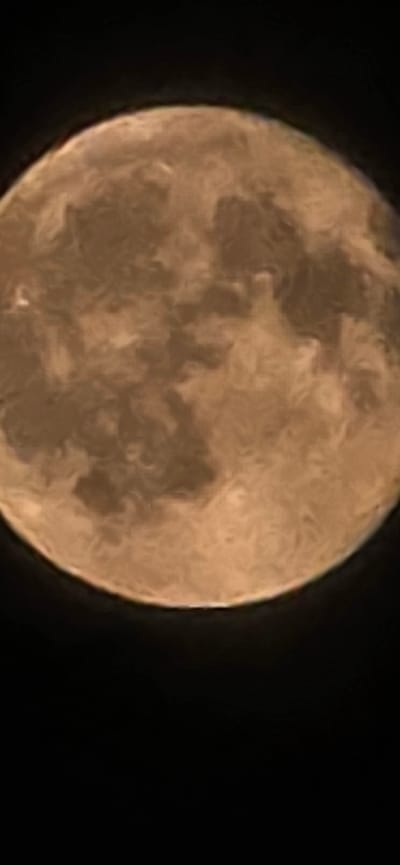 The image shows a close-up, slightly blurred view of the moon against a black background. The moon is predominantly tan and brown in color, with visible variations in shading that suggest surface features such as craters and maria. The overall impression is that of a full or nearly full moon. The black background implies the vastness of space, with no other celestial bodies or atmospheric phenomena clearly discernible. The image is tightly cropped, focusing solely on the moon. There is no indication of any urban environment, city, or geographical location such as Dzhalka, Russia.