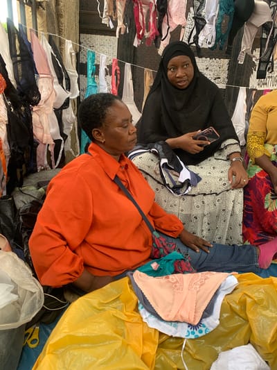 In Nyanya, Nigeria, two women are seated amidst a market stall filled with women's undergarments. The woman on the left, wearing an orange shirt, looks to her right. The woman on the right wears a black hijab and a patterned dress. She holds a mobile phone. Several bras are displayed hanging on a line behind them, and a pile of underwear is in the foreground. The environment suggests an outdoor market setting with a rough, textured background wall and various plastic sheeting.