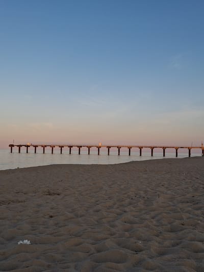 The image shows a long pier extending from the sandy beach into a calm body of water. The sky is a gradient of soft blue and pink, suggesting either sunrise or sunset. The pier is supported by numerous pilings and has railings along its length. There are small structures or buildings located on the pier. The sandy beach in the foreground exhibits ripples from the wind or tide. The overall scene is serene and depicts a coastal landscape in Shkorpilovtsi, Bulgaria.