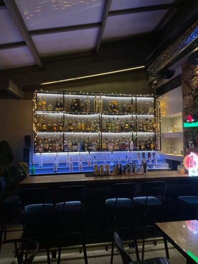 This interior shot captures a modern bar area, likely in a restaurant or pub, at night. The scene is devoid of people, suggesting a quiet moment either before opening, after closing, or during a lull.
The focal point is the illuminated bar counter and the extensive display of alcoholic beverages behind it. On the left, a large, industrial-style shelving unit made of metal mesh is filled with numerous bottles of wine and spirits. This section is adorned with warm white fairy lights, creating a sparkling effect. To its right, a sleek, multi-tiered display showcases an array of clear spirit bottles, dramatically lit from below by bright blue LED strip lighting. Above the bar, a rugged stone wall serves as a backdrop, featuring a glowing green Heineken star logo and a red and white circular sign with the brand name "CAMPARI" visible.
In front of the bar, a row of dark-colored bar stools with blue-grey upholstered seats are neatly arranged, awaiting patrons. To the far left, a large window or partition with frosted panels and dark framing is visible. A dark, reflective ventilation duct runs along the ceiling on the left side of the frame. A security camera is mounted on the wall near the top center. Some green plants are visible on the floor in the bottom right corner, adding a touch of natural element to the contemporary setting. The lighting throughout the space is dim, with the bar's illumination providing the main light sources, highlighting the extensive drink selection.