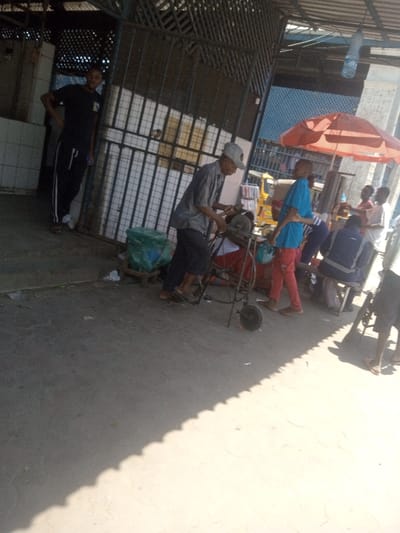 The image depicts an outdoor, semi-covered commercial area in Mombasa, Kenya. In the center, an older male individual, wearing a light grey baseball cap, a grey long-sleeved shirt, and dark trousers, operates a foot-pedal-driven grinding or sharpening machine mounted on a wheeled metal frame. He is bent forward, holding a metallic object against the machine's abrasive wheel. Adjacent to him, a younger male in a blue t-shirt and red trousers stands, observing the operation. To the left, a male in a dark t-shirt and dark track pants with white side stripes stands with his left hand on his hip, facing towards the central activity. To the right, under a large orange market umbrella, multiple individuals are gathered. One male, wearing a dark blue shirt, is seated on a low stool. Other figures are partially obscured, standing or sitting within this shaded zone. A light-colored auto-rickshaw (tuk-tuk) is visible in the background behind this group. The ground is paved concrete, marked by strong, distinct shadows cast from overhead structures, indicating direct sunlight. The environment includes metal grilles, a corrugated metal roof, and white-tiled wall sections. A clear plastic water bottle is suspended from the roof structure, and green plastic bags are positioned on a low platform near the sharpening station.
