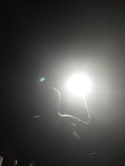 A silhouette of a person is visible against a bright light source, which appears to be a spotlight or a large lamp. The person's left arm is raised, and they are facing away from the camera. A faint green rectangular light is visible to the left of the person's head. The background is dark, suggesting it is nighttime or a dimly lit indoor space. The overall impression is dramatic due to the strong contrast between the dark silhouette and the bright light. The location in the city of Il'mino, Russia, is not discernible from the image itself.