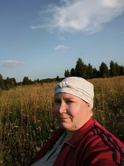A selfie-style, medium-angle, full-body shot captures a woman from the chest up, standing in a sunlit meadow with a dense line of trees in the background. The woman is positioned on the right side of the frame, facing left and looking towards the camera with a slight smile. She is wearing a red zippered jacket with black stripes on the sleeves and a light-colored headscarf patterned with green leaves and light blue flowers. Her skin is fair, and she has a mole on her left jawline. The meadow is filled with tall, dry grass and scattered wildflowers. The sky above is a clear blue with a few wispy white clouds. The scene appears to be an outdoor, natural setting, possibly on the outskirts of the village of Zipunovo, Russia.