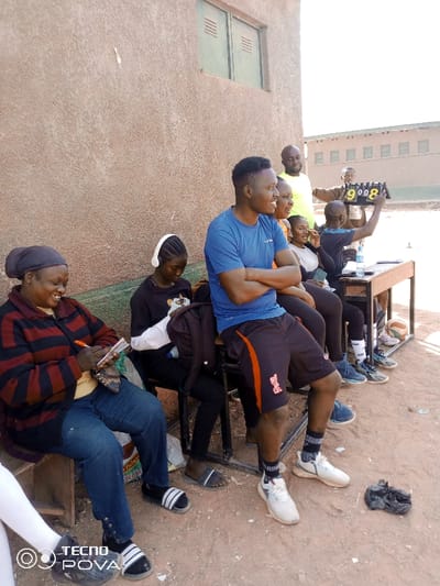 The image presents an outdoor scene in Kaduna South, Nigeria, featuring eight individuals under clear daylight conditions. The background consists of a light brown, stucco-textured building wall with two green louvered windows on the upper left. The ground is reddish-brown soil.
From left to right:
A dark-skinned woman, wearing a dark headscarf, a red and black striped long-sleeved top, and blue jeans, is seated on a wooden bench. She is smiling and actively writing on a small pad of paper with an orange marker. Next to her, a dark-skinned young female wearing white over-ear headphones, a dark long-sleeved top, and dark trousers is seated on a chair, looking downwards. A black backpack rests against her chair.
Centrally, a dark-skinned man with a short haircut is seated on a chair, wearing a blue t-shirt with a white logo, black shorts with an orange accent, white socks, and white sneakers. He is smiling and looking towards the right, with his arms crossed. Behind him, a partially visible dark-skinned woman in a light top and a bald dark-skinned man in a bright yellow-green t-shirt are present. To their right, another dark-skinned woman in a dark top is visible.
Further right, a dark-skinned man in a brown shirt stands behind a plain wooden table, holding up a black flip-style scoreboard that displays the numbers "9008." Another dark-skinned man in a blue shirt is seated at the table. A plastic water bottle is on the table, and a partially deflated basketball rests on the ground underneath it. A crumpled black plastic bag is visible on the ground near the central man's feet. The overall scene suggests a gathering or event, possibly sports-related, with individuals observing or participating in an official capacity.