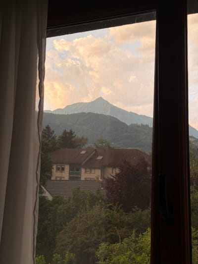 The image shows a view through a window, likely from a residential building in Cluses, France, at sunset. A light-colored curtain hangs on the left side of the frame. The window pane has a fine mesh screen. In the foreground, lush green trees and foliage are visible, obscuring parts of houses with brown tiled roofs. A two-story building with a light-colored facade and multiple windows is partially visible among the trees. In the middle ground, layers of forested mountains recede into the distance. The sky is filled with dramatic clouds illuminated by the warm, soft light of the setting sun, casting an orange and pink glow. The distant mountain peaks are silhouetted against the sky. The overall scene conveys a sense of peaceful domesticity against a backdrop of natural grandeur.