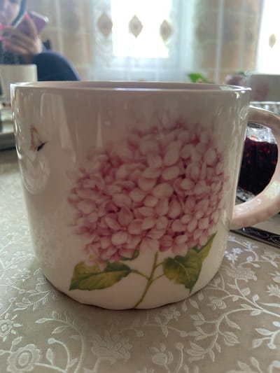 A close-up, eye-level shot features a ceramic mug adorned with a pink hydrangea design. The mug is primarily white with a light pink handle. To the right of the mug, partially visible, is a glass jar containing a dark red substance, likely jam or preserves. The mug and jar are placed on a textured, light-colored tablecloth with an embossed floral pattern. In the background, a blurred figure is seen holding a pink smartphone. The lighting suggests an indoor setting, possibly near a window, indicated by the soft, diffused light and the presence of what appears to be window blinds in the upper background. The scene appears to be a still life, focusing on the decorative mug and its contents. The location within the city of Askarovo, Russia, is not discernible from the image itself, as there are no geographical markers or contextual clues.