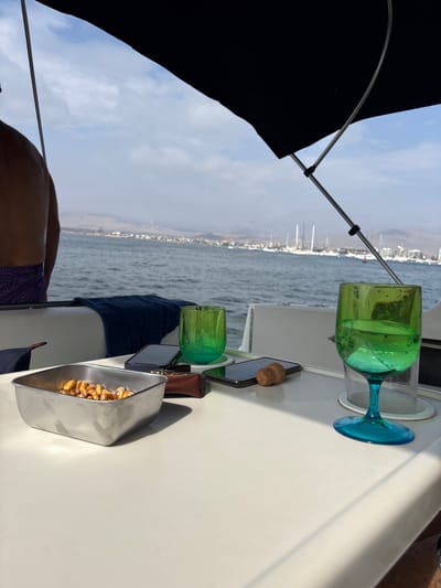 The image depicts a view from a boat, with the cityscape of Ancón, Peru, visible in the background across the water. The foreground features a white surface upon which sits a tall glass filled with a green liquid and a blue stem, a phone, a wine cork, a brown leather item, a smaller glass filled with green liquid, a blue towel, and a metal container of nuts. A person wearing purple swimwear is partially visible in the lower left. A black umbrella provides shade.
