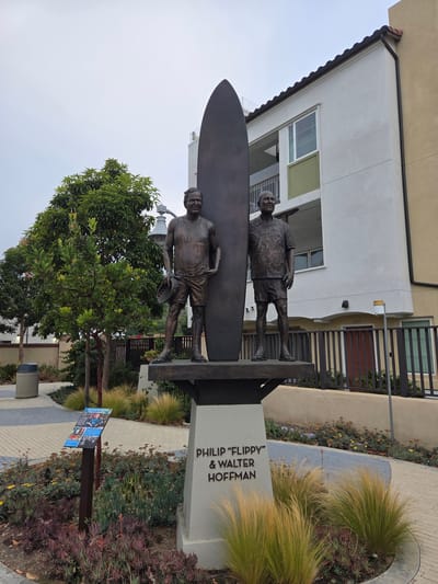 The image shows a bronze statue of two men, Philip "Flippy" and Walter Hoffman, standing next to a surfboard. The statue is located outdoors in Dana Point, California, USA. The men are depicted in casual attire, with one wearing a tank top and shorts and the other a patterned short-sleeved shirt and shorts. Both are wearing sandals. The statue is mounted on a pedestal with the names of the individuals inscribed on it. In the background, there is a modern building with a light-colored facade, balconies, and a tiled roof. A black metal fence runs along the front of the building. The foreground features landscaping with various plants, including ornamental grasses and succulents, and a paved walkway with a decorative pattern. A trash receptacle is visible on the left side of the image, and a sign with information about the individuals is placed near the statue. The sky is overcast.