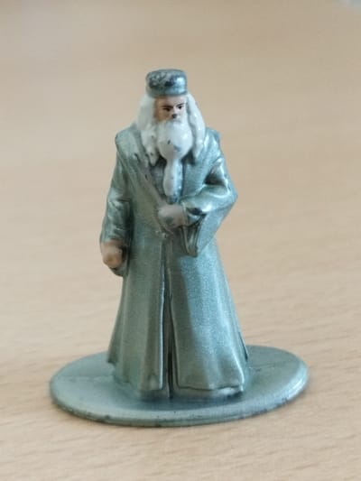 This media file is a close-up, eye-level photograph of a miniature figurine of a man, likely a character from a fantasy or historical setting, standing on a circular base. The figurine is predominantly a muted, metallic blue-green color, with lighter silver accents on his hat and robe. He has a long, white beard and mustache, and his face is painted with subtle details for eyes, nose, and mouth. He is wearing a hat and a flowing, floor-length robe with what appears to be a tie or sash around his neck. His left hand is tucked into his robe, and his right hand is slightly visible in front of him. The figurine is positioned on a flat, light brown surface, which is likely a wooden table, with a subtle wood grain visible. The background is a plain, out-of-focus beige. There is no visible text in the image. The lighting suggests an indoor setting, likely during the daytime, with no specific weather cues. The figurine itself is the sole focus, and no activity or event is depicted beyond its static pose. The overall impression is that of a collectible or toy.