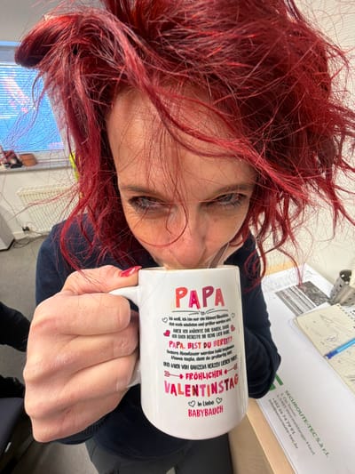 The image displays an individual with voluminous, dyed red hair, seen in a close-up as they consume a beverage from a white ceramic mug. The subject's eyes are partially visible beneath the hair and are directed towards the camera. The left hand, adorned with red fingernail polish on the thumb and index finger, securely holds the mug. The mug's exterior features a multi-colored German inscription, prominently displaying "PAPA," "FRÖHLICHEN VALENTINSTAG," and concluding with "In Liebe Babybauch." The background reveals an indoor setting, identified as an office in Grevenmacher, Luxembourg, supported by a visible document on a desk to the right. This document displays the name "SECUROUTE-TEC S.à r.l." and the address "L-6701 Grevenmacher, Luxembourg." The environment further includes a window with horizontal blinds, a white radiator, and various office items on the desk surface, such as a metal mesh pen holder, a blue pen, and additional papers.