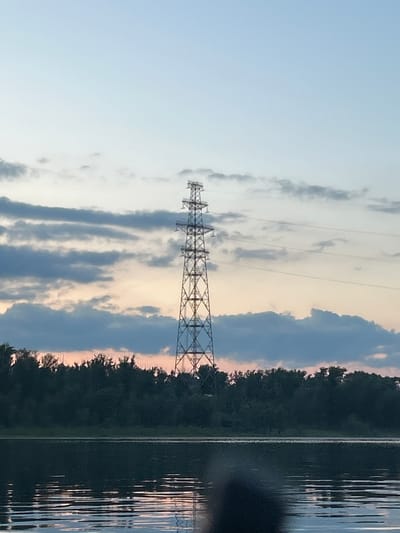 An image depicts a tall, lattice-like electricity pylon situated in the distance, positioned centrally in the frame. The pylon stands against a sky filled with a gradient of colors, transitioning from a pale blue at the top to soft orange and pink hues closer to the horizon. Scattered clouds, varying in shade from light gray to darker, denser formations, dot the sky. Below the sky, a dense line of dark green trees forms a tree line, creating a stark contrast against the lighter sky. In the foreground, a body of water reflects the subtle colors of the sky and the dark silhouettes of the trees. The water's surface shows ripples, suggesting a gentle movement. The scene appears to be captured during twilight or dawn. The location is identified as Ol'khovskoye, Russia. No people or specific actions are discernible in the image.