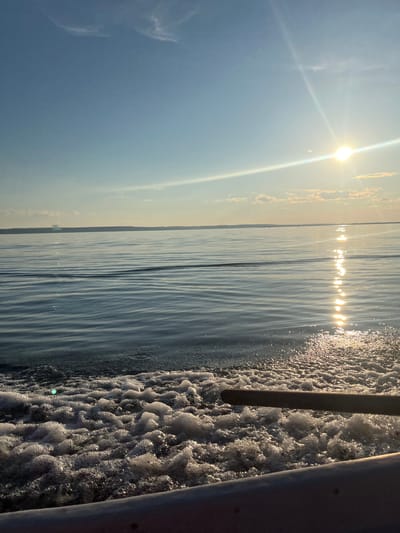 This image depicts a serene scene of a body of water, likely a lake or river, with a clear sky. The sun is prominently featured, casting a bright glare and reflecting on the water's surface, creating a shimmering effect. The water appears calm, with gentle ripples indicating a possible breeze. On the right side of the image, the wake of a boat is visible, characterized by foamy, disturbed water and two metallic poles, suggesting the perspective is from on board a vessel. The sky is a soft blue with a few wispy clouds. The time of day appears to be either early morning or late afternoon due to the low angle of the sun and the warm, golden light. The overall mood is peaceful and tranquil, evoking a sense of calm and relaxation. The location is identified as Siukterka, Russia, which could be a specific village, town, or natural area within Russia. There is no visible text in the image.
