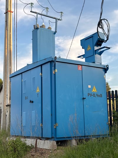 The image shows a large blue electrical transformer box situated outdoors, likely in a utility area. The transformer box is tall and rectangular, with several doors and vents on its sides. A smaller, tiered blue structure with a junction box containing coiled cables is mounted on top of the main box. Electrical insulators and wires are visible connecting to the top of the transformer and extending towards a concrete utility pole on the left side of the frame. A yellow triangular warning symbol with a lightning bolt is visible on the front of the transformer. Cyrillic text, including "РУ-О,ЧКВ", is also present on the box. The transformer is surrounded by tall grass and dry weeds, with a black metal fence visible on the right. The sky above is partly cloudy. There are no people visible in the image. The setting appears to be in a rural or semi-urban area, possibly in the city of Chigiri, Russia, given the presence of Cyrillic text and the style of the utility equipment.