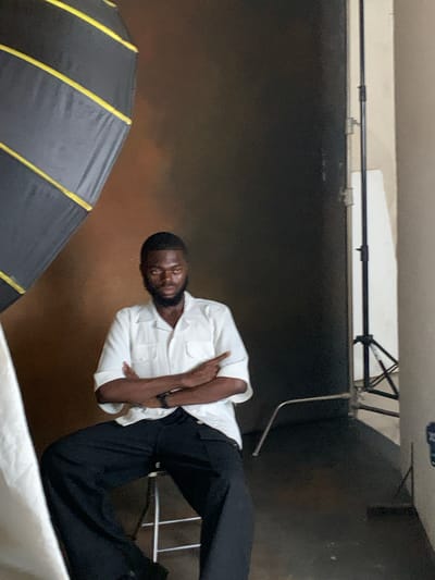 The image depicts a young Black man seated on a stool, facing the camera. He is wearing a white short-sleeved collared shirt with two pockets on the chest and black wide-leg pants. His arms are crossed, and his expression is serious.

The setting appears to be a photography studio. To the left of the man, a large, dark softbox with yellow accents is visible, positioned at an angle. Behind him, the backdrop is a dark, textured brown. To the right, a studio light stand with a white reflector or backdrop is present. The floor is dark.

The image does not contain any information that would allow for its location to be specified in the city of Barikin Jirga Summa, Nigeria.