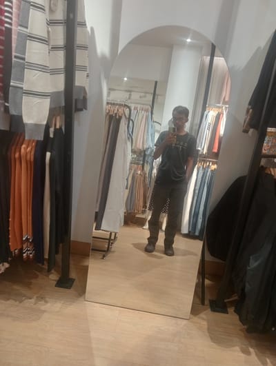 A person is taking a selfie in a full-length mirror inside a clothing store. The person, who appears to be a young man, is wearing a dark t-shirt and dark pants. He has glasses and short hair. He is holding up his phone to take the picture, with his left hand in his pocket.

The store setting is visible in the reflection of the mirror. There are racks of clothing on either side of the person. On the left, there are several sweaters and cardigans hanging on hangers, in colors like cream, gray, orange, and navy. On the right, more clothing is visible, including what appear to be dresses and tops. The floor is made of light-colored wood planks. The lighting appears to be artificial, suggesting the photo was taken indoors. There are no clear indicators of the time of day or weather outside. The overall mood seems casual and relaxed, as the person is casually posing for a photo in a retail environment.

The t-shirt the person is wearing has some text on it, but it is reversed in the mirror and difficult to read clearly. It appears to be in a light color on the dark fabric.