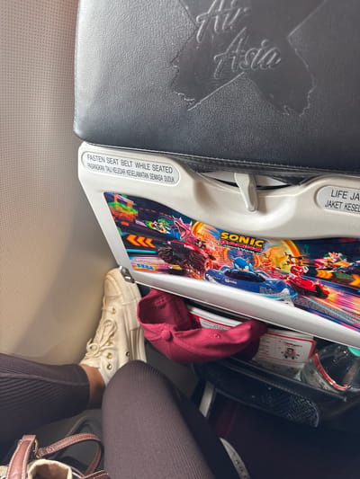 The image shows the interior of what appears to be an airplane cabin. The seat in front has a tray table folded down featuring "Sonic Racing" promotional graphics. The tray table has safety instructions in English and Malay. The seat is likely on an AirAsia flight, based on the logo visible on the headrest. Visible under the tray table are a red cap, a pack of cards, and a half-empty plastic bottle. A person is seated, wearing brown pants and white sneakers. The person is likely traveling on AirAsia, with the image taken on or near the city of Sepang, Malaysia, which is where the Kuala Lumpur International Airport is located.