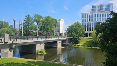 The image shows a bright, sunny day in Linköping, Sweden, with a clear blue sky and a few wispy clouds. The scene features a wide concrete bridge spanning a calm, dark green body of water. The bridge has black railings and decorative hanging baskets filled with red and purple flowers. Two modern office buildings are visible on the far bank of the water. The building on the left is white with a red "Scandic" logo. The building on the right is a taller, multi-faceted glass and white structure with "Danske Bank" and "AW" logos visible. Lush green trees and grass surround the buildings and line the banks of the water, creating a serene urban landscape. There are no people or animals clearly visible in the image.