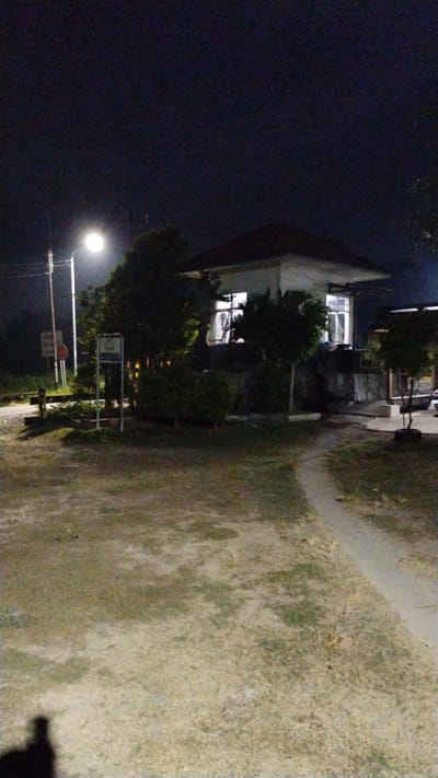 A vertical, low-angle, nighttime exterior shot captures a small, brightly lit building with a red-tiled roof. The building appears to be a guardhouse or office, with large windows illuminating the interior. A streetlamp on the left casts a bright glow on the surrounding trees and a dirt path that winds through a grassy area in the foreground. The ground is covered in dry, patchy grass with some exposed soil. A shadow of a person is visible in the lower-left corner of the image. The background is dark, suggesting it is late at night. Signs are visible near the streetlamp.