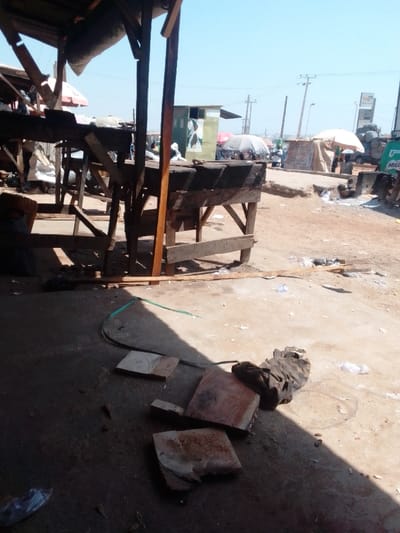 The image displays an outdoor market scene under a clear blue sky, located in the city of 0801712591, Nigeria. The immediate foreground consists of a partially shaded concrete or paved surface, transitioning to a sunlit dirt ground in the mid-ground. Several empty, dark brown wooden market stalls, constructed from rough planks and lacking goods, occupy the left and central foreground. One stall features a dark fabric covering.
Scattered debris is visible on the ground, including a coiled green hose, several pieces of light-colored, irregularly shaped material (possibly broken tiles or wood), a crumpled dark fabric object, and various small plastic items. In the mid-ground, a light-green shack with a white graphic is positioned to the left. Further back, numerous indistinct figures of people are present amidst additional market stalls, some covered by large umbrellas (one prominent grey) and tarps. Utility poles with overhead power lines traverse the scene horizontally. On the far right, a partial view of a vehicle, likely a truck, shows the text "MILL" on its side, adjacent to a white tarp-covered structure. A large, partially visible billboard or sign is in the distant background.