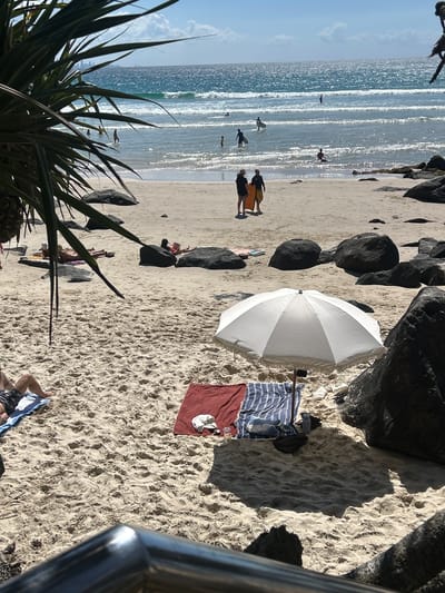 This photograph depicts a scene on a beach in Coolangatta, Australia. The image is oriented sideways. The ocean is visible on the left, with surfers in the water. On the right, the sandy beach contains large rocks, a white umbrella, and several towels including one of red. People are lying on the beach and standing near the water. A plant is visible in the lower left corner, with a railing on the lower right.