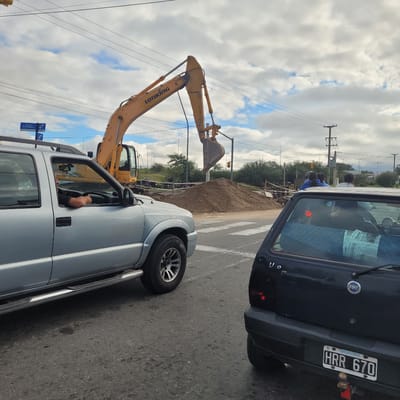 The image depicts a construction scene on a street in what appears to be San Fernando del Valle de Catamarca, Argentina, based on the license plate visible on a black Fiat Uno. A yellow LONKING excavator is positioned near a pile of dirt on a pedestrian crosswalk. A silver pickup truck is to the right of the frame with an unidentified driver inside. A group of individuals wearing blue attire and hard hats are observed in the background behind the pile of dirt. Power lines and street signs including an Avenida del Bicentenario sign are visible in the scene.